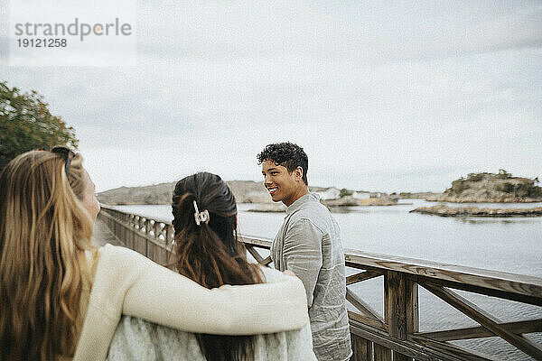 Lächelnder junger Mann mit Freundinnen  die auf einer Brücke in der Nähe eines Sees spazieren gehen