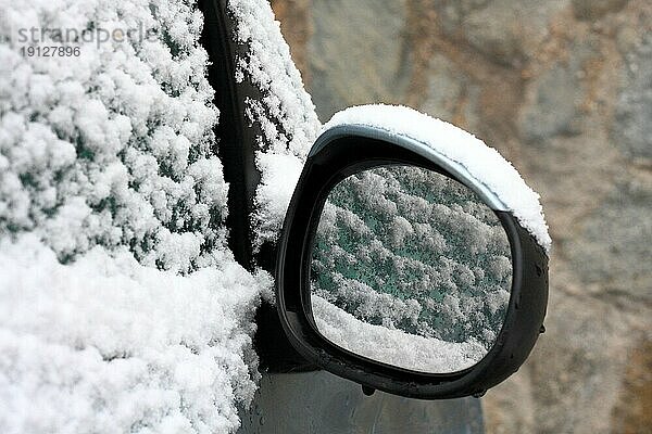 Eingeschneites Auto (Ausschnitt) mit Seitenspiegel  in dem sich Schnee spiegelt  Hintergrund Hauswand in Unschärfe