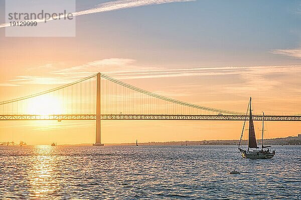 Blick auf die Brücke 25 de Abril  berühmtes touristisches Wahrzeichen von Lissabon  die Lisboa und Almada über den Fluss Tejo verbindet  mit der Silhouette einer Touristenyacht bei Sonnenuntergang. Lissabon  Portugal  Europa