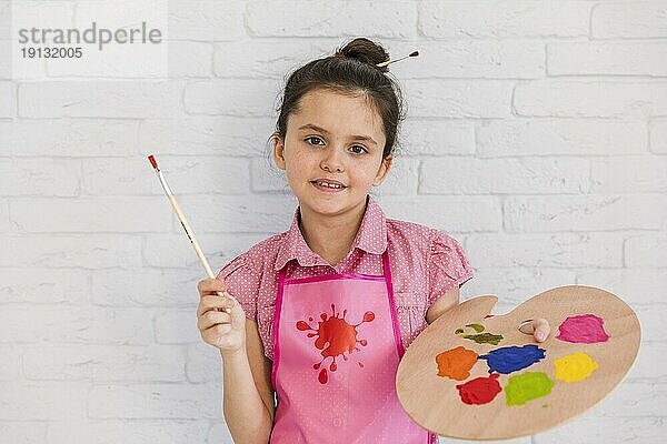 Lächelnde Porträt Mädchen stehen gegen weiße Backsteinmauer hält Pinsel bunte Palette