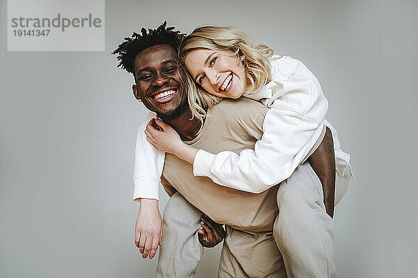 Cheerful young man piggybacking girlfriend against gray background