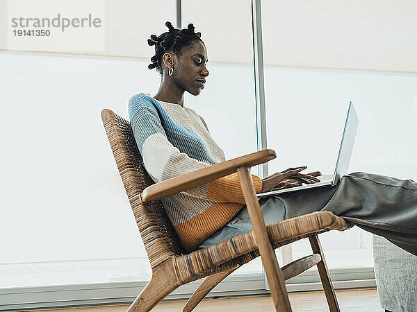 Junge Frau sitzt mit Laptop im Sessel am Fenster zu Hause
