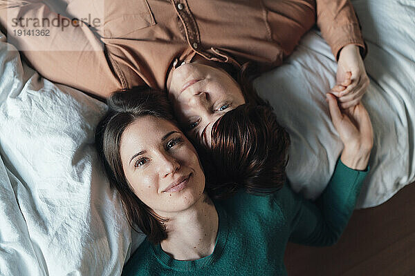 Smiling lesbian couple holding hands and lying down at home