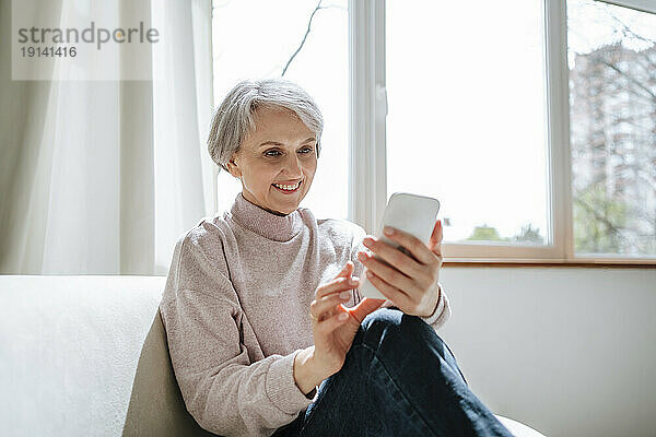 Lächelnde reife Frau  die am Fenster sitzt und ihr Smartphone benutzt