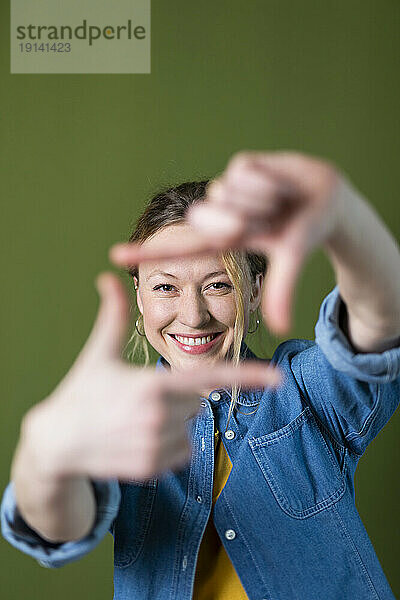 Smiling young woman making finger frame in front of green wall