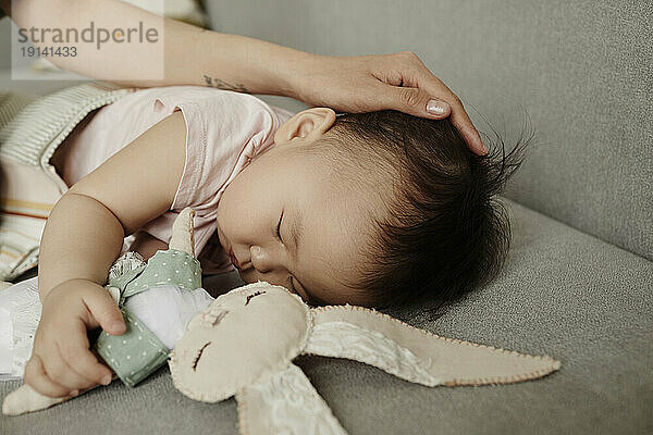 Mother stroking daughter sleeping with stuffed toy on sofa at home