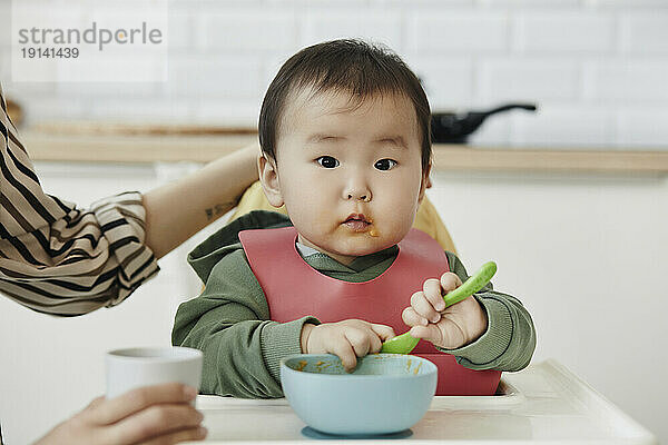Cute daughter with bowl and spoon sitting in high chair at home