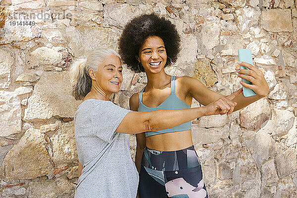 Lächelnde Frau macht ein Selfie mit ihrer Mutter vor einer Steinmauer