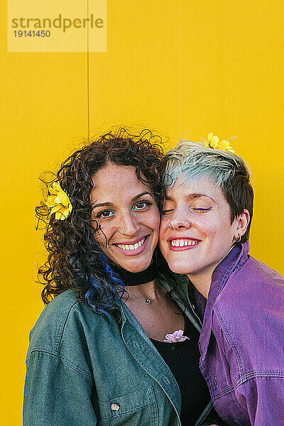 Smiling woman by lesbian friend with flowers on hair in front of yellow wall