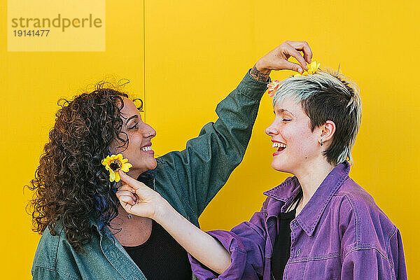 Lesbisches Paar legt sich vor gelber Wand gegenseitig Blumen ins Haar