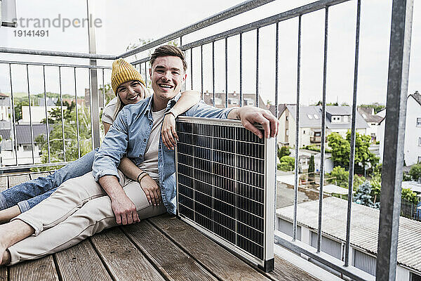 Glückliches junges Paar sitzt mit Solarpanel am Geländer auf dem Balkon