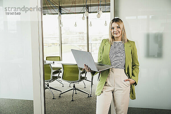 Selbstbewusste junge Geschäftsfrau hält Laptop neben Glastür im Büro