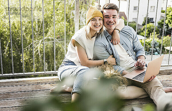 Happy couple sitting with laptop and dog on balcony