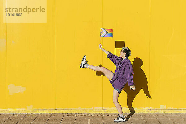 Lesbische Frau tanzt mit einem Bein und hält eine Regenbogenfahne vor einer gelben Wand