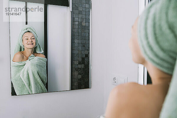 Lächelnde Frau  in ein Handtuch gehüllt  schaut in den Badezimmerspiegel