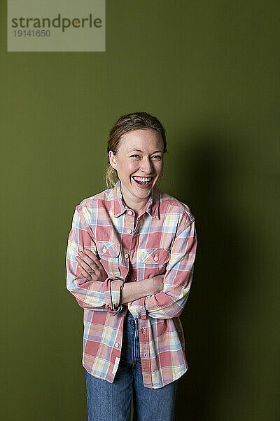 Young woman in plaid shirt laughing with arms crossed in front of green wall