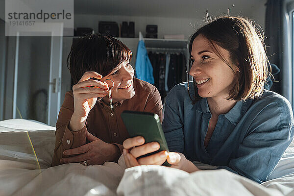 Lächelnde Frau schaut lesbische Freundin an  die zu Hause ihr Smartphone hält