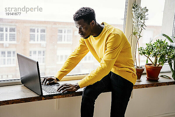 Konzentrierter Mann mit Laptop  der zu Hause auf der Fensterbank sitzt
