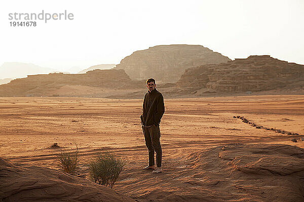 Junger Mann mit Händen in den Taschen steht bei Sonnenuntergang in der Wüste