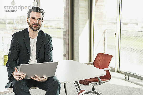 Selbstbewusster Geschäftsmann sitzt mit Laptop am Tisch im Büro