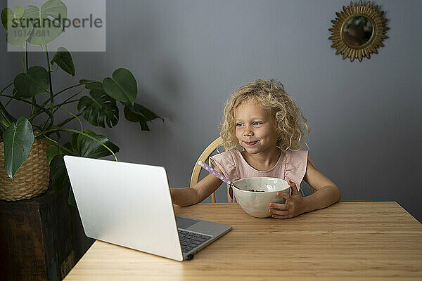 Smiling blond girl using laptop and having breakfast at dining table