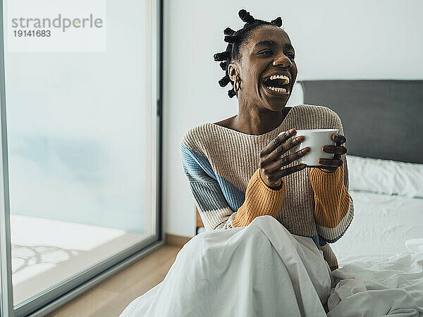 Laughing woman holding cup of coffee sitting on bed
