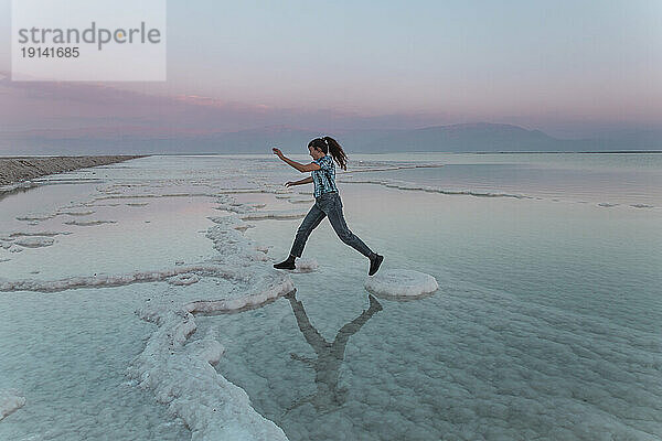 Junge Frau springt bei Sonnenuntergang auf Salzformationen im Toten Meer