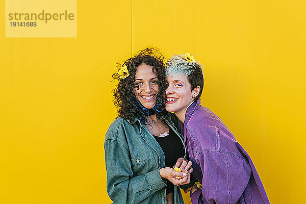 Happy lesbian couple with flowers on hair standing in front of yellow wall