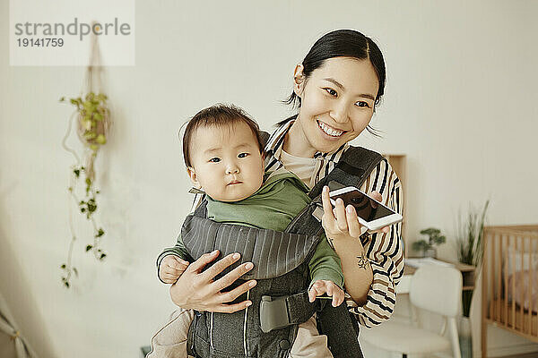 Happy woman taking through speaker phone carrying daughter in baby carrier at home