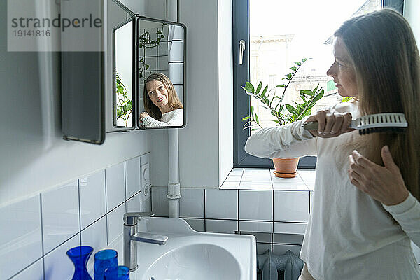 Happy mature woman brushing hair in bathroom at home