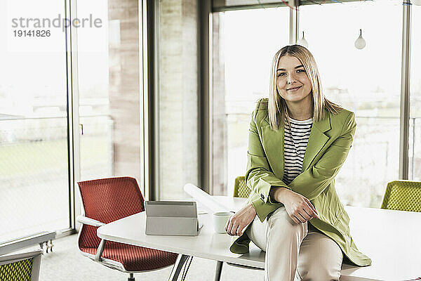 Selbstbewusst lächelnde Geschäftsfrau sitzt mit Tablet-PC am Tisch im Büro