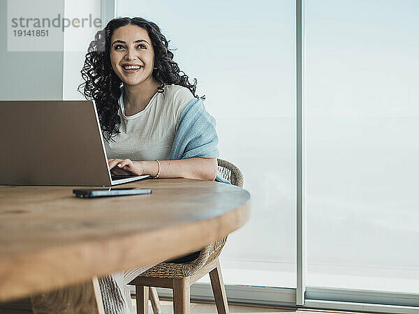 Smiling young woman using laptop looking away sitting in chair at home