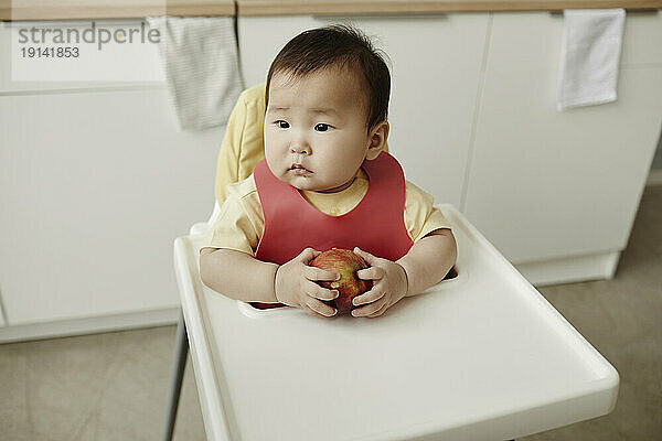 Cute girl holding apple sitting on high chair at home