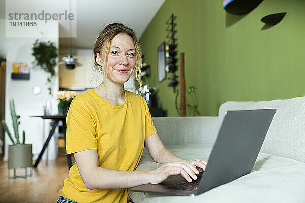 Junger Freiberufler im gelben T-Shirt arbeitet zu Hause mit Laptop