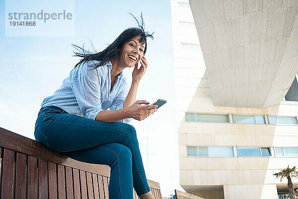 Glückliche Geschäftsfrau hält Smartphone auf Bank sitzend