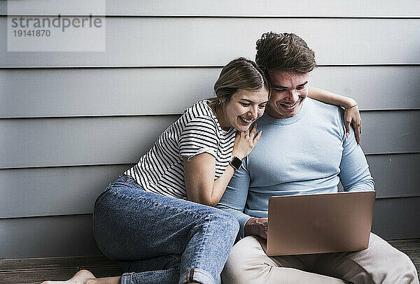 Glückliches junges Paar sitzt vor der Wand und benutzt Laptop
