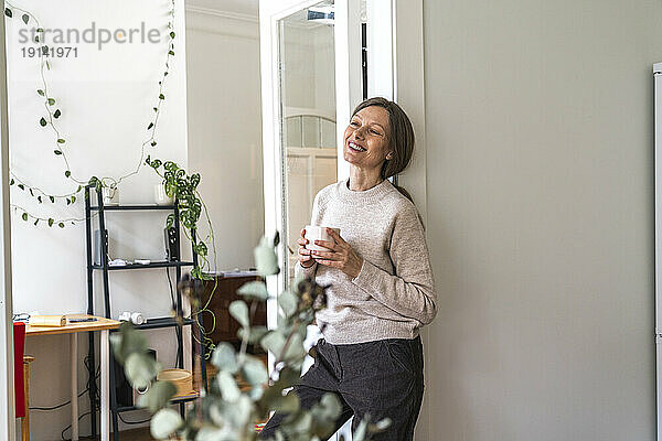 Glückliche reife Frau lehnt mit Kaffeetasse an der Wand