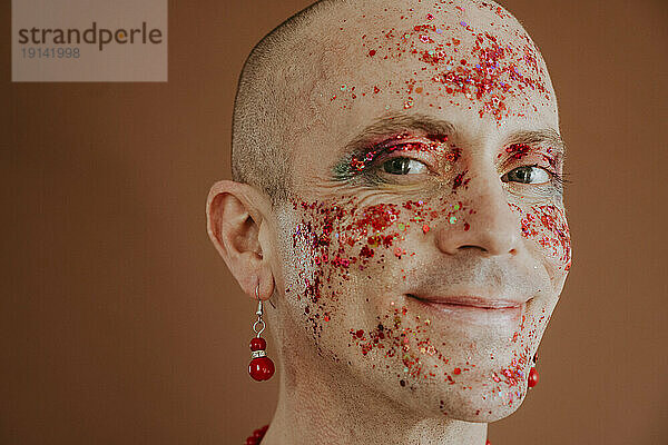 Smiling face of drag queen with red glitter on face against brown background