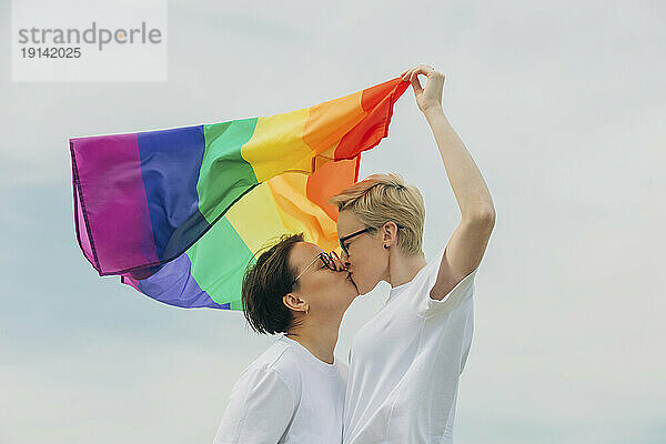 Liebevolle Lesben mit Regenbogenfahne küssen sich unter dem Himmel