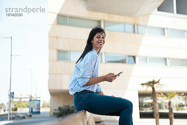 Glückliche Geschäftsfrau mit Smartphone sitzt auf Bank