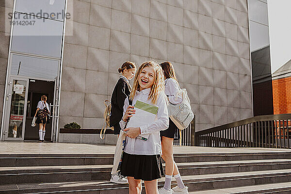 Glückliches Mädchen  das vor dem Schulgebäude steht  mit Freunden im Hintergrund