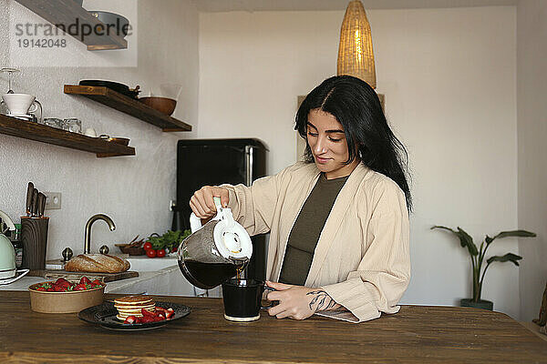 Junge Frau gießt frisch gebrühten Kaffee in eine Tasse am Küchentisch