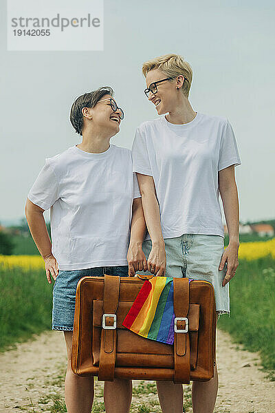 Fröhliche Lesben halten einen Koffer mit Regenbogenfahne in der Hand