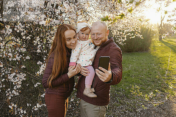 Vater und Mutter machen ein Selfie mit ihrer Tochter  die neben einem Baum im Park steht