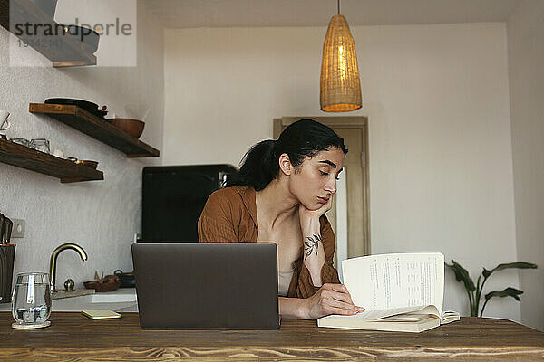 Junge Frau lernt zu Hause mit Buch und Laptop auf dem Tisch