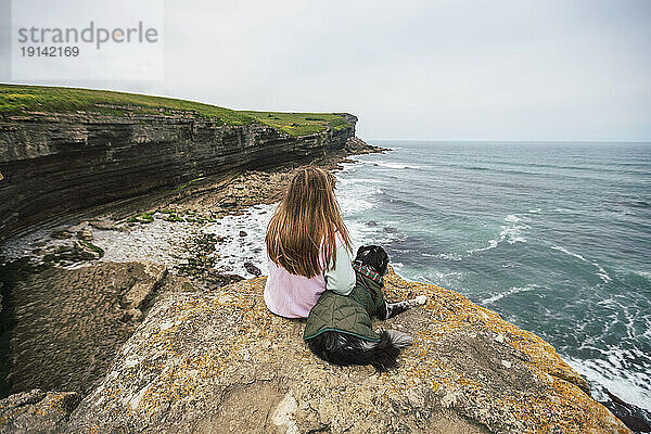 Junge Frau sitzt mit Hund auf einem Felsen und schaut aufs Meer