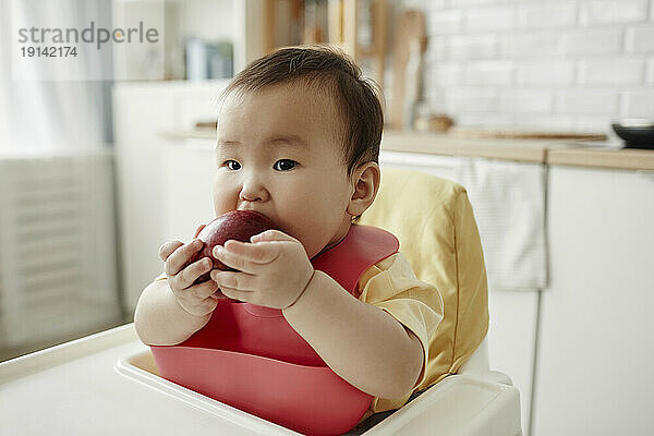 Nettes Mädchen  das Apfel isst und zu Hause auf dem Kindersitz sitzt
