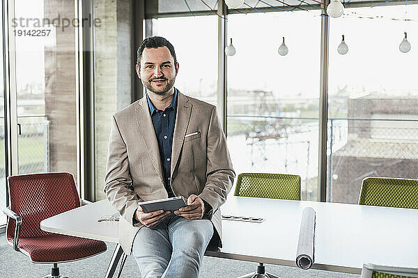 Selbstbewusster Geschäftsmann mit Tablet-PC sitzt am Tisch im Büro
