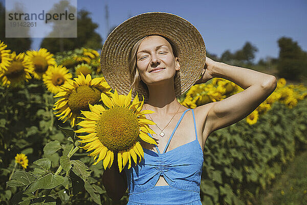 Lächelnde Frau mit Sonnenhut hält Sonnenblume im Feld