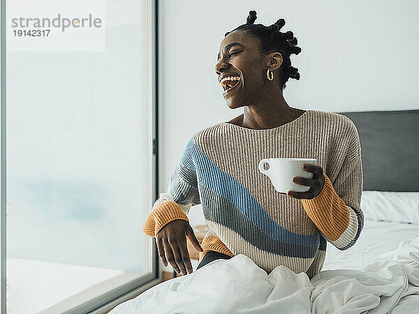 Woman laughing and holding coffee cup sitting on bed at home